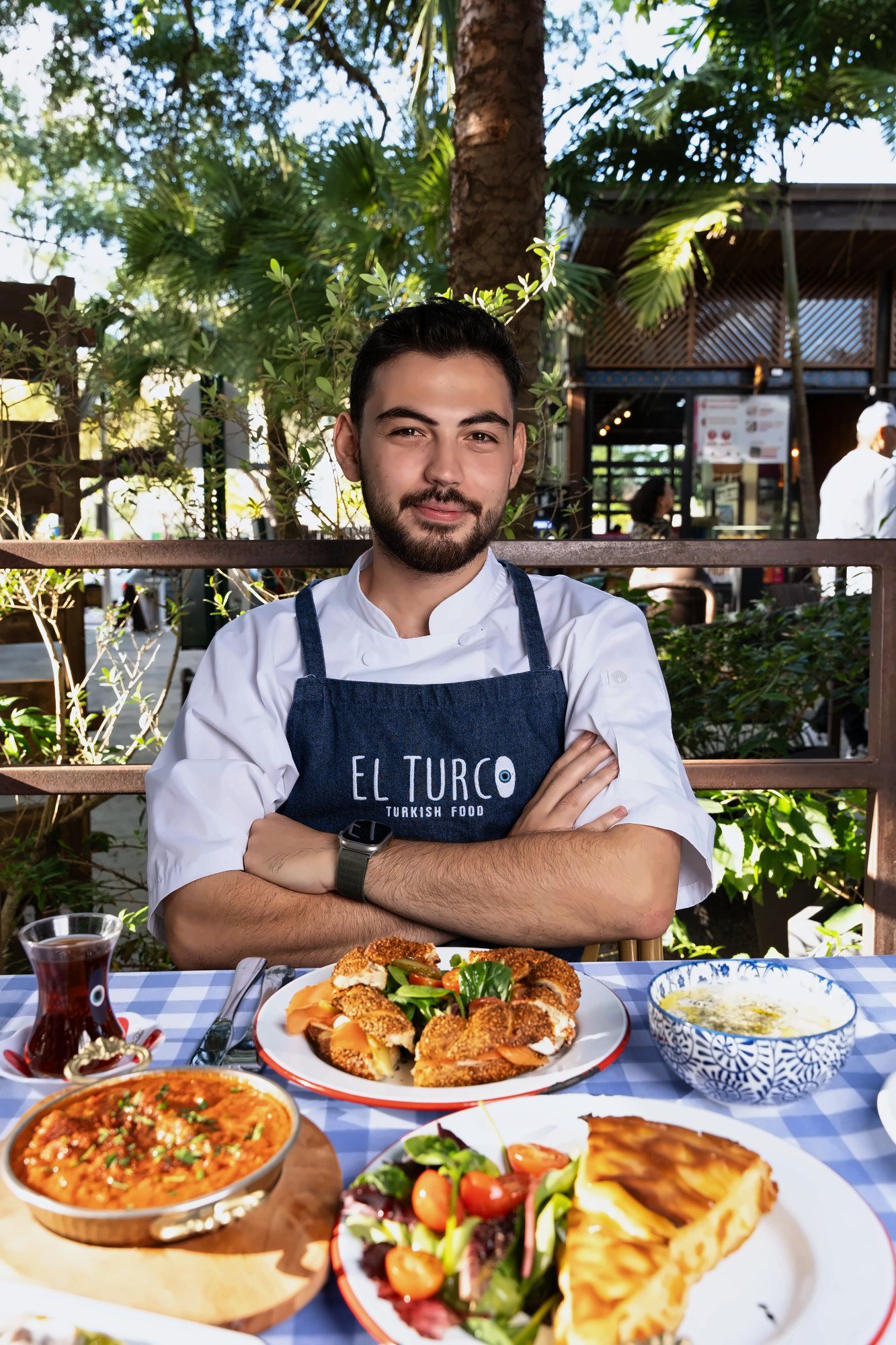 El Turco chef with arms crossed behind a table of Turkish dishes on a leafy patio